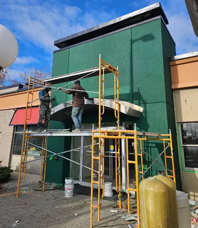 Two crew members on scaffolding reinstalling a metal canopy over a storefront entrance freshly finished in dark green stucco